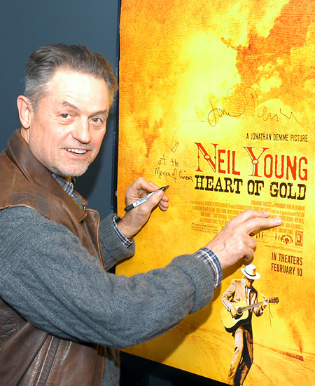 Jonathan Demme looking at camera, smiling and holding pen to sign a yellow poster that reads Neil Young Heart of Gold