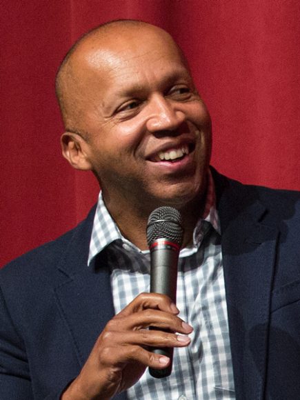 Bryan Stevenson wearing checkered button down shirt, smiling, holding a microphone on red background