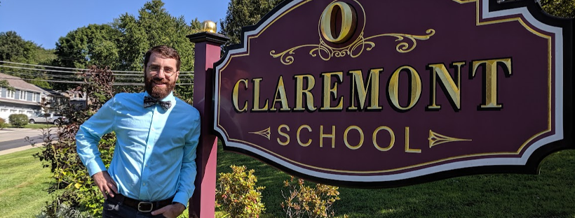 Photograph of a white man in a blue shirt and bow tie standing aside a sign for the Claremont School
