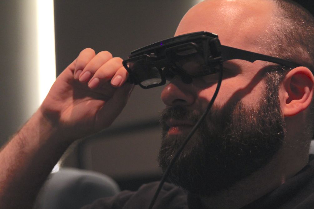 man wearing closed captioning glasses with one hand holding front side of frames in theater
